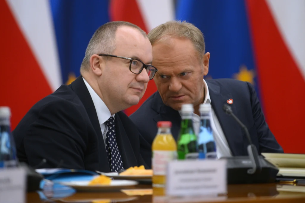 Adam Bodnar i Donald Tusk Dwaj mężczyźni w garniturach siedzą przy stole konferencyjnym, prowadząc rozmowę wśród butelek z napojami oraz dokumentów, w tle widoczne flagi Polski i Unii Europejskiej.