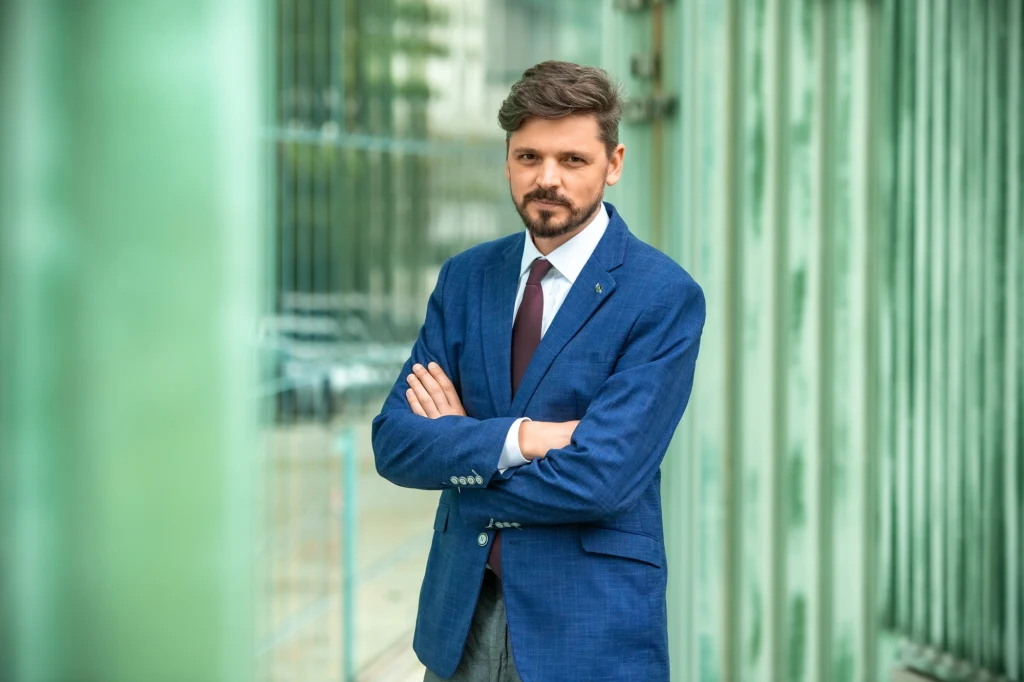 Bartosz Pilitowski, prezes Fundacji Court Watch Polska, która od lat monitoruje funkcjonowanie sądów w Polsce Mężczyzna w granatowej marynarce i koszuli z krawatem stoi z założonymi rękami na tle przeszklonej nowoczesnej architektury.