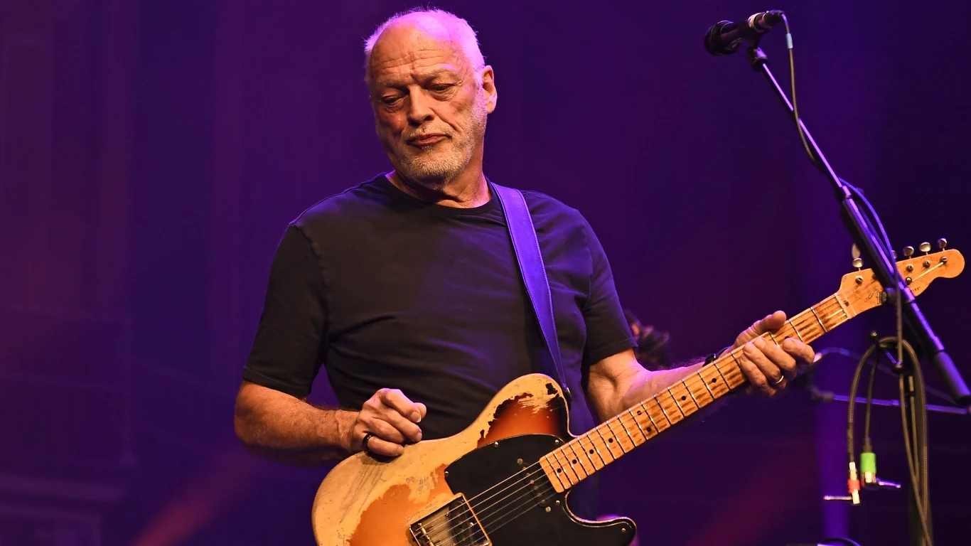 David Gilmour Mężczyzna z siwą brodą gra na gitarze elektrycznej na scenie oświetlonej kolorowymi światłami, ubrany w czarną koszulkę, w tle mikrofon i ciemna scenografia.