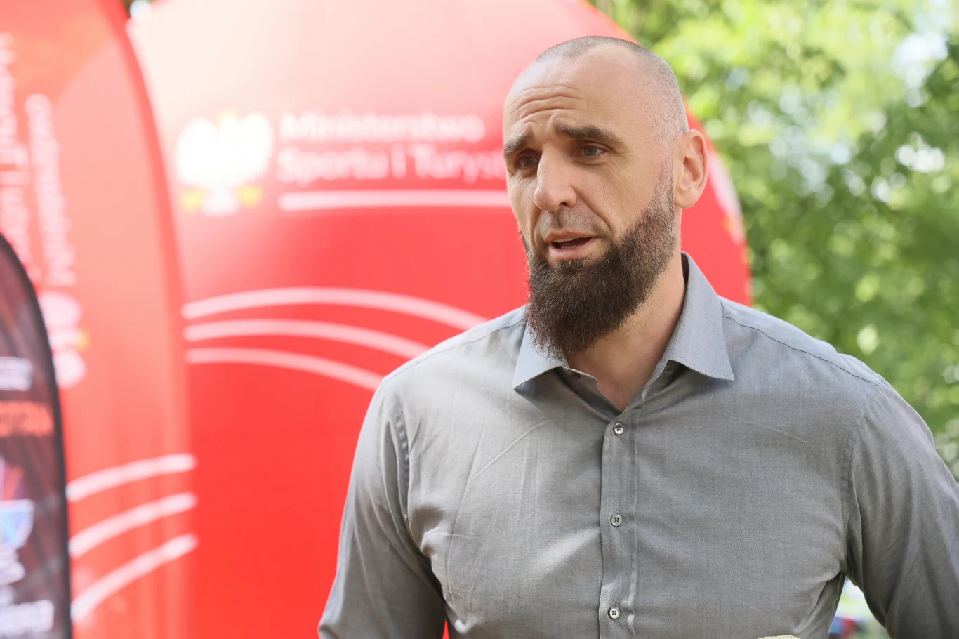 Marcin Gortat Mężczyzna z krótkimi włosami i gęstą brodą w szarej koszuli stoi na tle czerwonej konstrukcji z logo Ministerstwa Sportu i Turystyki, w otoczeniu zieleni.