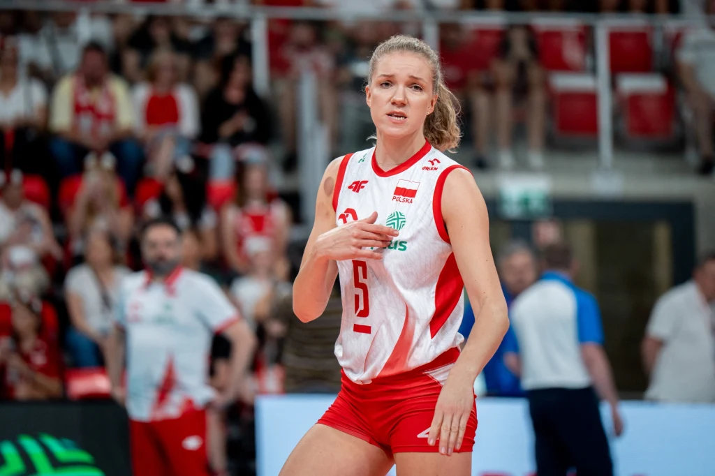 Siatkarka w stroju reprezentacji Polski stoi na boisku podczas meczu, w tle widoczni inni zawodnicy oraz kibice na trybunach.