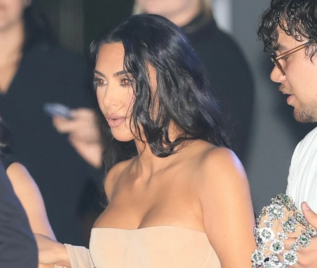 Kim Kardashian skupiła na sobie uwagę w ciekawy sposób Kobieta o długich, ciemnych włosach ubrana w beżową, dopasowaną sukienkę, stoi obok mężczyzny z okularami trzymającego błyszczący przedmiot ozdobiony kamieniami. W tle widoczne są inne osoby, lekko rozmazane.
