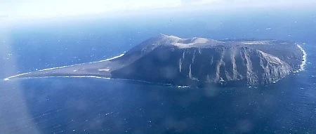 Surtsey w 1999 r. Wyspa wulkaniczna otoczona lazurową wodą oceanu, o nierównym, stromym zboczu i wyraźnie widocznych ciemnych skałach.
