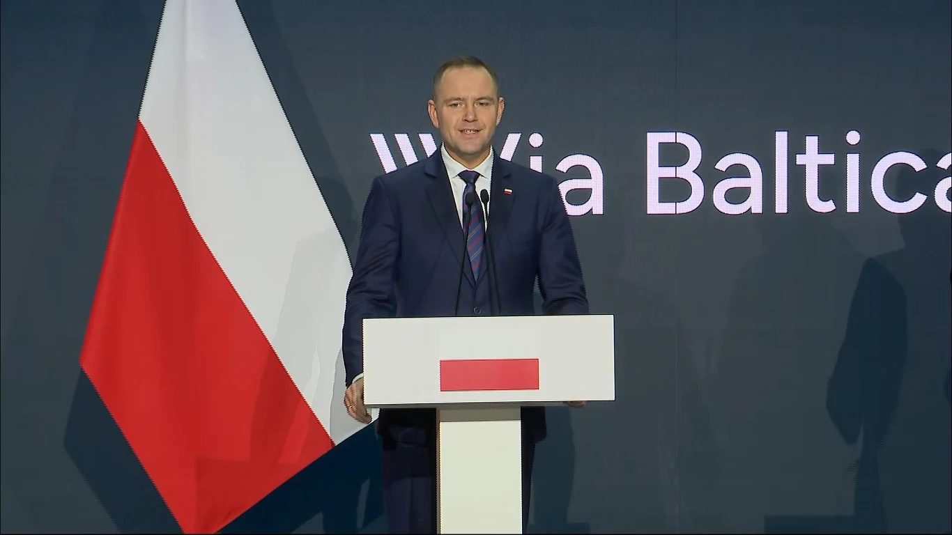 Mężczyzna w garniturze stoi przy mównicy na tle dużej polskiej flagi oraz napisu Via Baltica, wydarzenie odbywa się w oficjalnej atmosferze.