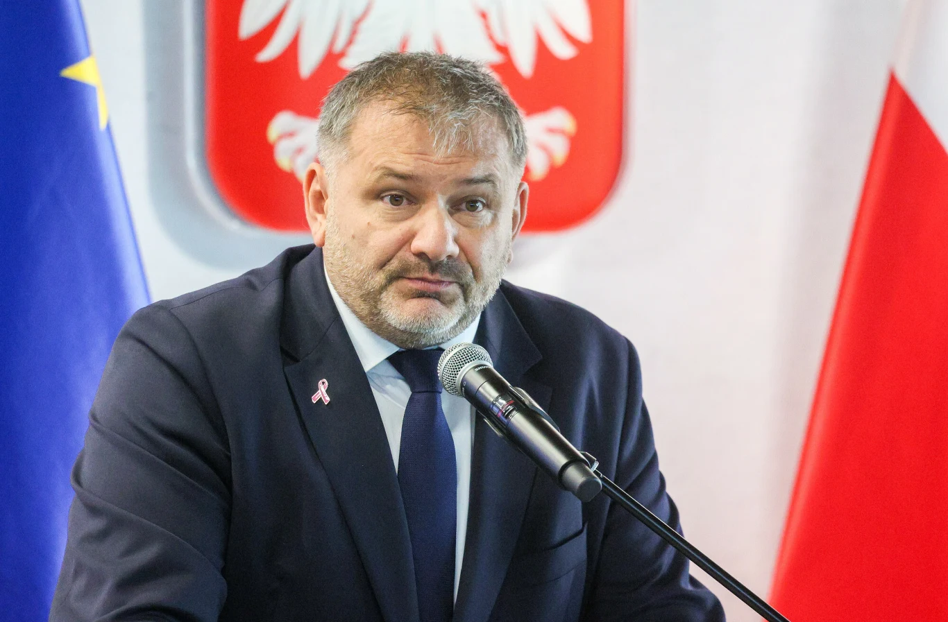 Minister sprawiedliwości i prokurator generalny Waldemar Żurek Mężczyzna w garniturze przemawiający do mikrofonu, w tle flaga Unii Europejskiej, flaga Polski oraz herb Polski.