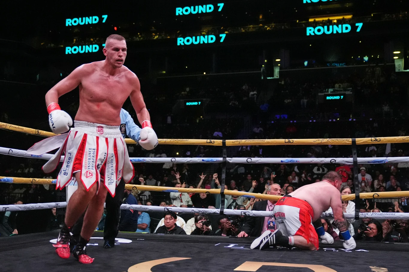 Dwóch bokserów podczas walki w ringu, jeden stoi z podniesioną głową, drugi klęczy przy linach, publiczność ogląda pojedynek, na tablicach wyświetlany jest siódmy runda.