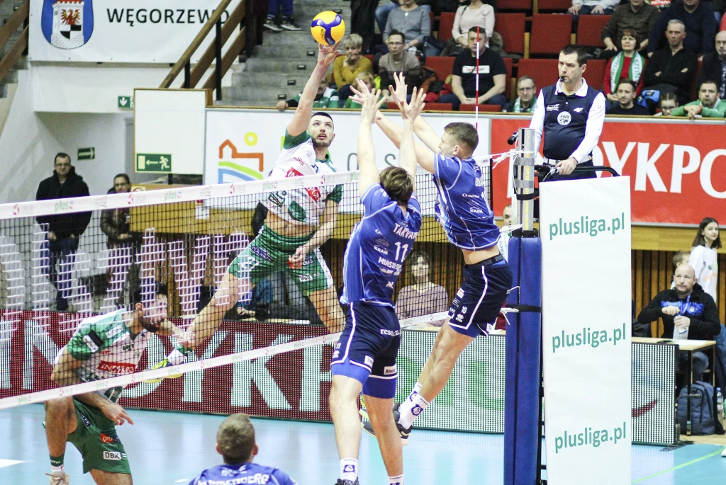 Intensywna akcja podczas meczu siatkówki, zawodnik drużyny w zielono-białym stroju wykonuje atak, próbując przebić piłkę ponad blokującymi graczami drużyny w niebieskich strojach, widownia kibiców obserwuje wydarzenie z trybun.