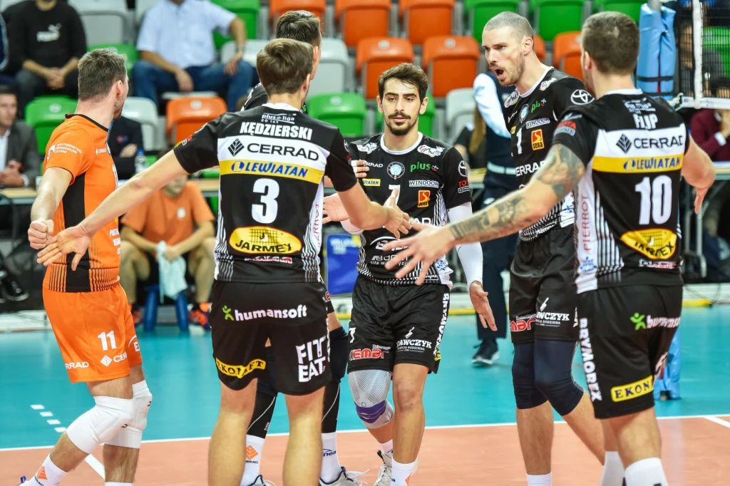 Grupa siatkarzy podczas meczu, zawodnicy ubrani w stroje sportowe, w tle trybuny z kibicami i kolorowe krzesła, atmosfera skupienia i współpracy na boisku