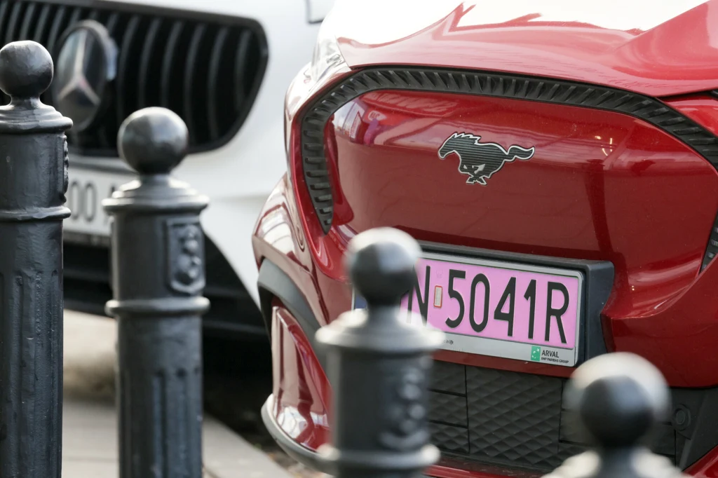 Fragment przodu czerwonego samochodu marki Ford Mustang Mach-E z różową tablicą rejestracyjną obok białego auta marki Mercedes, na pierwszym planie metalowe słupki chodnikowe.