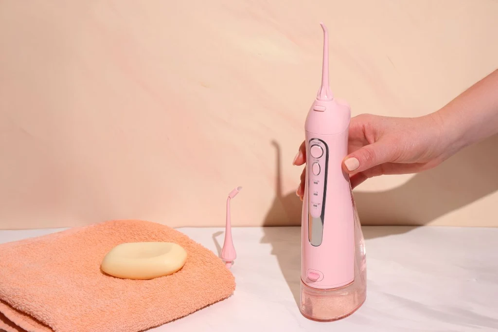 Na rynku dostępnych jest wiele opcji. Zacznij od najprostszego irygatora - efekty cię zaskoczą Ręka trzyma różowy irygator do jamy ustnej ustawiony na białej powierzchni obok pomarańczowego ręcznika i mydła, w tle jasna ściana.