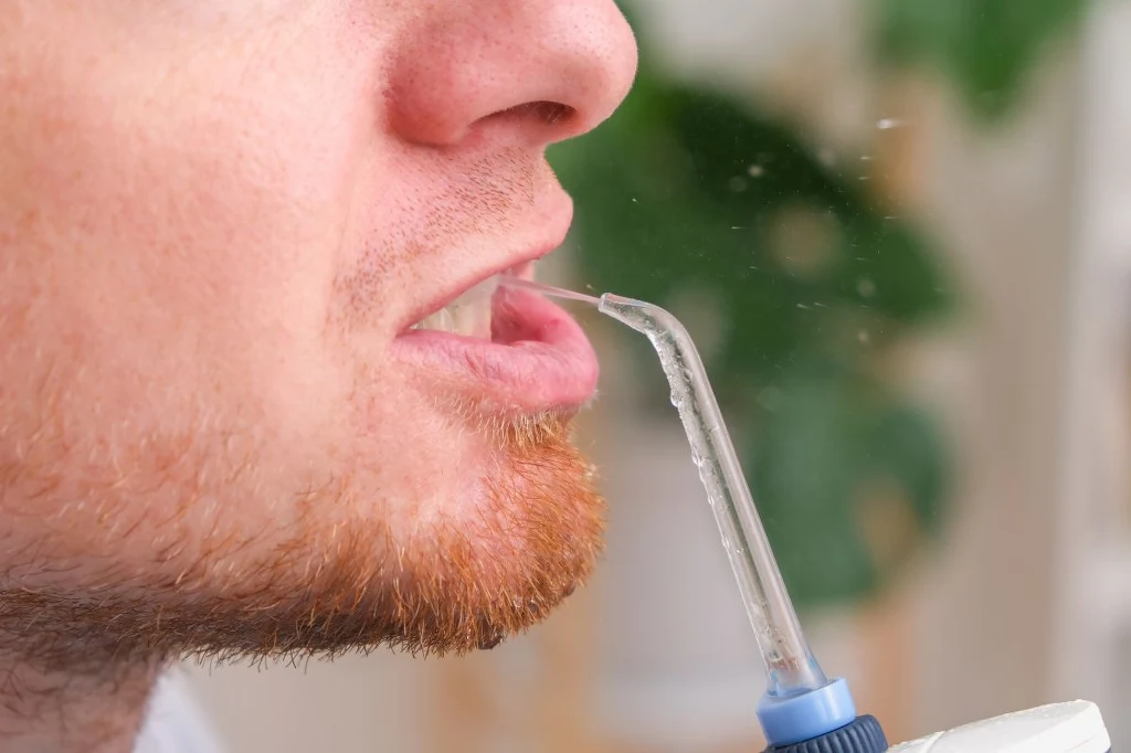 Z irygatora wypyszczany jest cienki strumień wody pod ciśnieniem, który skutecznie doczyszcza przestrzenie międzyzębowe Mężczyzna używa irygatora do czyszczenia przestrzeni międzyzębowych, z ust wydobywa się strumień wody.