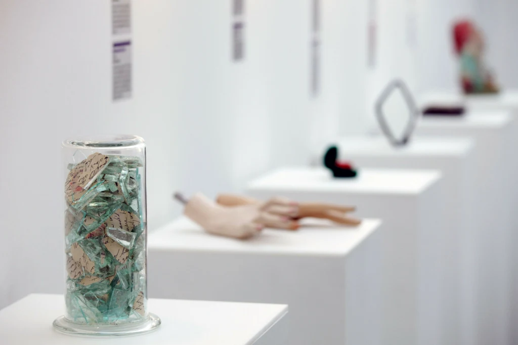 Wystawa w Muzeum Zerwanych Związków w Zagrzebiu Kilka ekspozycji artystycznych ustawionych na białych postumentach w galerii, na pierwszym planie szklany pojemnik wypełniony kawałkami szkła i papieru, w tle widoczne inne obiekty, w tym manekiny dłoni i geometryczne przedmioty.