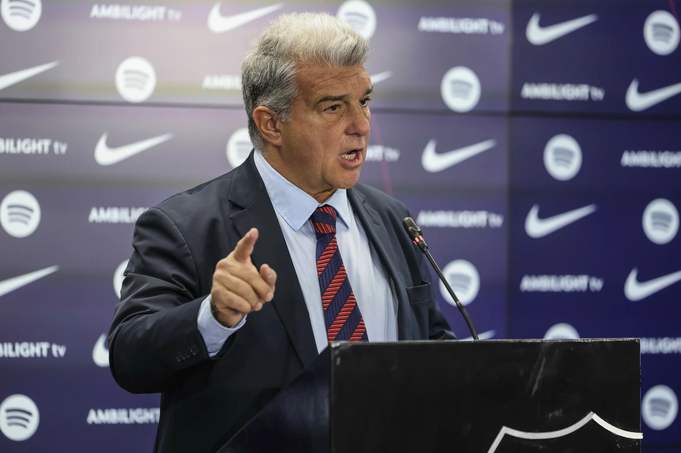 Joan Laporta, 14.01.2025 r. Mężczyzna w garniturze z szaro-siwych włosach przemawia na konferencji prasowej, gestykulując dłonią przy mównicy na tle ściany z logotypami sponsorów i klubu sportowego.