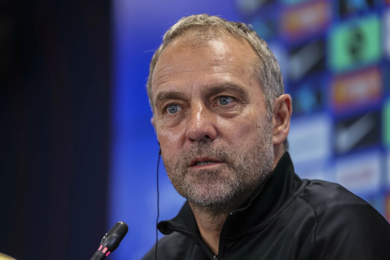 Mężczyzna z krótkimi, jasnymi włosami i zarostem, ubrany w czarną kurtkę, siedzi przed mikrofonem podczas konferencji prasowej. W tle rozmyte kolorowe tablice sponsorów sportowych.