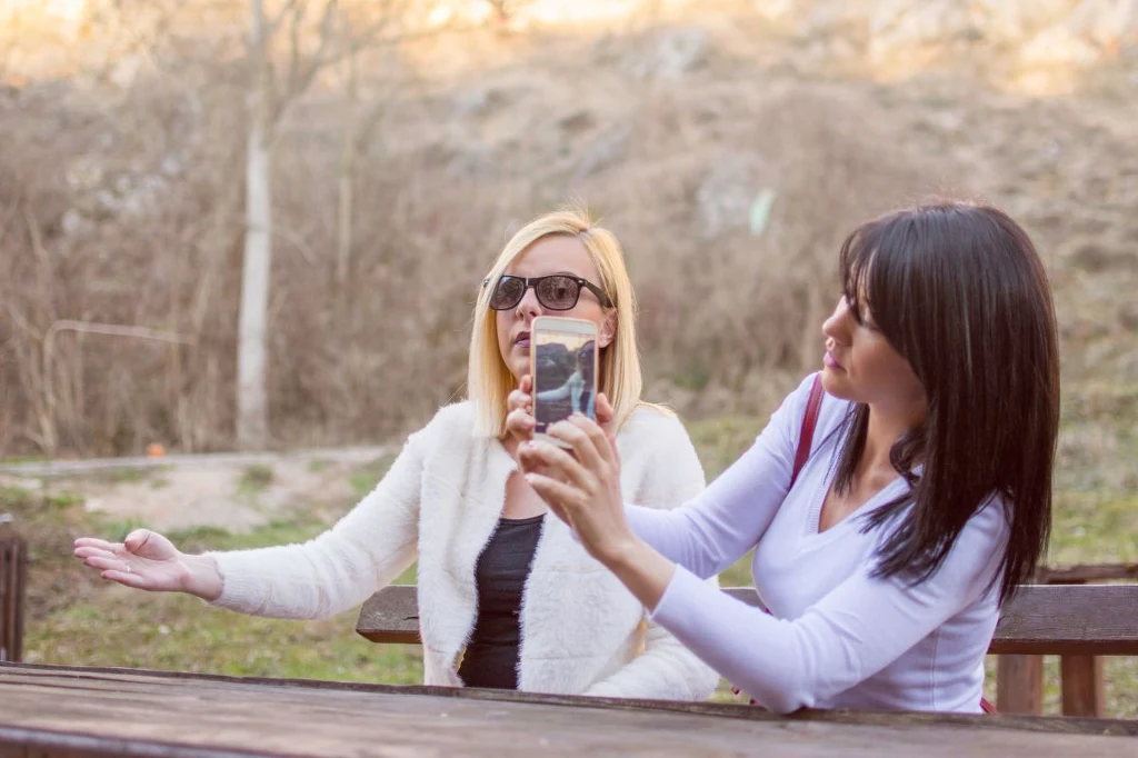 Dwie kobiety siedzące na drewnianej ławce w parku, jedna z nich nosi okulary przeciwsłoneczne, druga trzyma smartfona i wykonuje zdjęcie lub selfie. Otoczenie naturalne, w tle widoczne drzewa i sucha trawa.