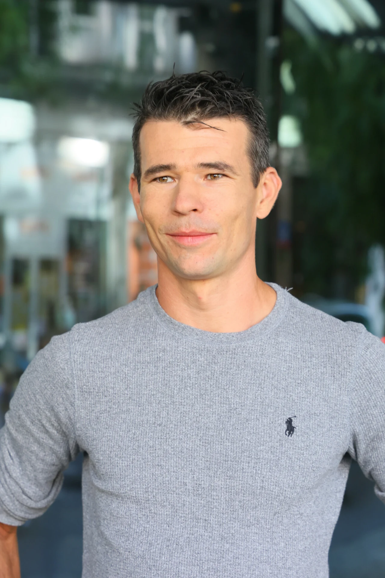 Adrian Kostera Uśmiechnięty mężczyzna o krótkich, ciemnych włosach, ubrany w szary sweter z drobnym logo, stoi w lekko rozmytym, miejskim otoczeniu.