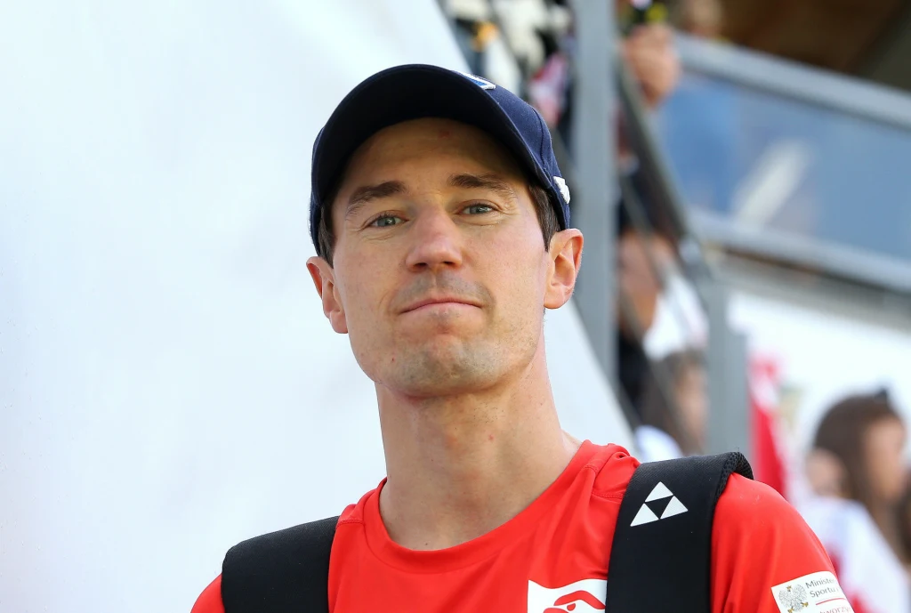 Kamil Stoch Mężczyzna w czerwonej koszulce sportowej i czapce z daszkiem, z plecakiem na ramionach, stoi na tle rozmytego tłumu podczas wydarzenia sportowego.