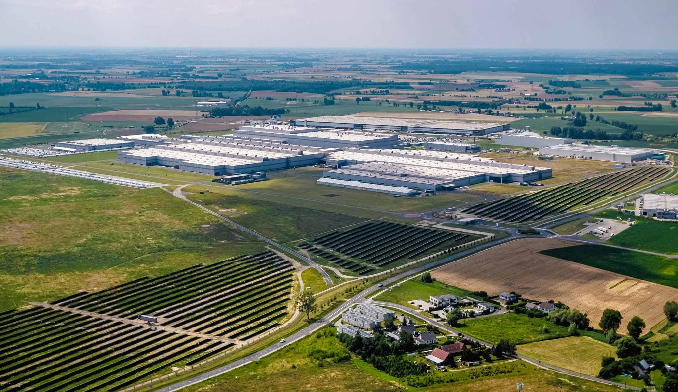 Zakład Volkswagena we Wrześni i farma fotowoltaiczna Rozległy kompleks przemysłowy otoczony polami uprawnymi i farmą fotowoltaiczną, liczne zabudowania w tle, droga dojazdowa prowadząca do fabryki, w oddali widoczna spokojna wiejska okolica.