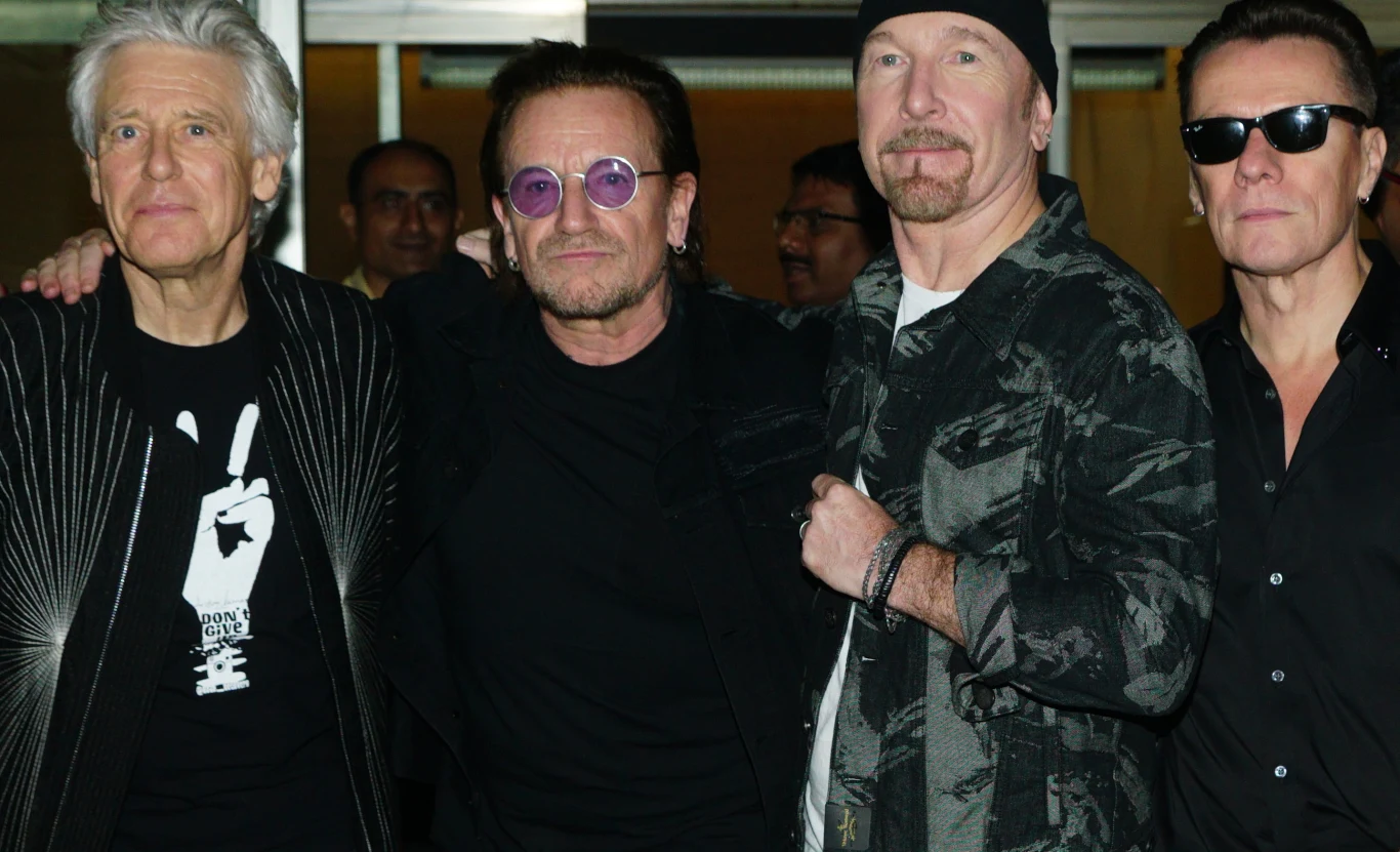 Zespół U2 wydał album "Boy" 45 lat temu Czterech mężczyzn pozuje razem ramiona na ramionach, wszyscy ubrani w ciemne ubrania, dwóch z nich nosi okulary przeciwsłoneczne, jeden ma bandanę na głowie, stoją blisko siebie na tle neutralnego wnętrza.