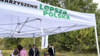Pod namiotem z logo Lepsza Polska grupa osób uczestniczy w warsztatach lub zajęciach plenerowych, siedząc przy stole z różnymi materiałami artystycznymi; w tle drzewa i otwarta przestrzeń.