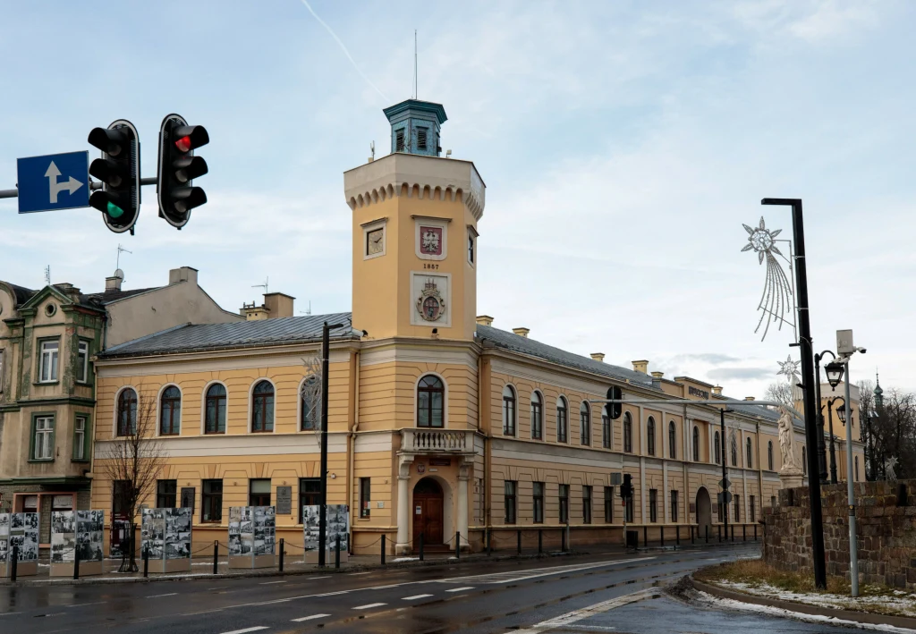 Historyczny budynek ratusza z wieżą zegarową, utrzymany w jasnych kolorach, położony przy głównej ulicy, z widocznymi lampami miejskimi i sygnalizacją świetlną, w tle ozdoba świąteczna w kształcie gwiazdy.