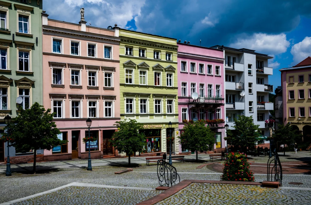 Kolorowe kamienice w centralnej części miasta, ozdobione balkonami i delikatnymi detalami architektonicznymi, pod nimi sklepy oraz lokalne punkty usługowe. Przed budynkami rozpościera się zadbany plac z kostką brukową, stylowymi latarniami, małymi drze...