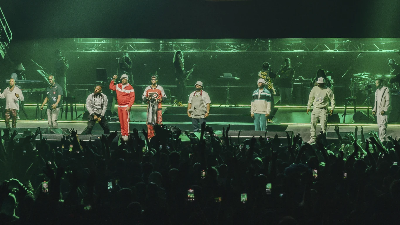 Wu-Tang Clan Grupa muzyków i wykonawców w kolorowych ubraniach stoi na scenie podczas koncertu, tłum ludzi pod sceną unosi ręce i nagrywa wydarzenie telefonami, intensywne zielone oświetlenie tworzy dynamiczną atmosferę występu.