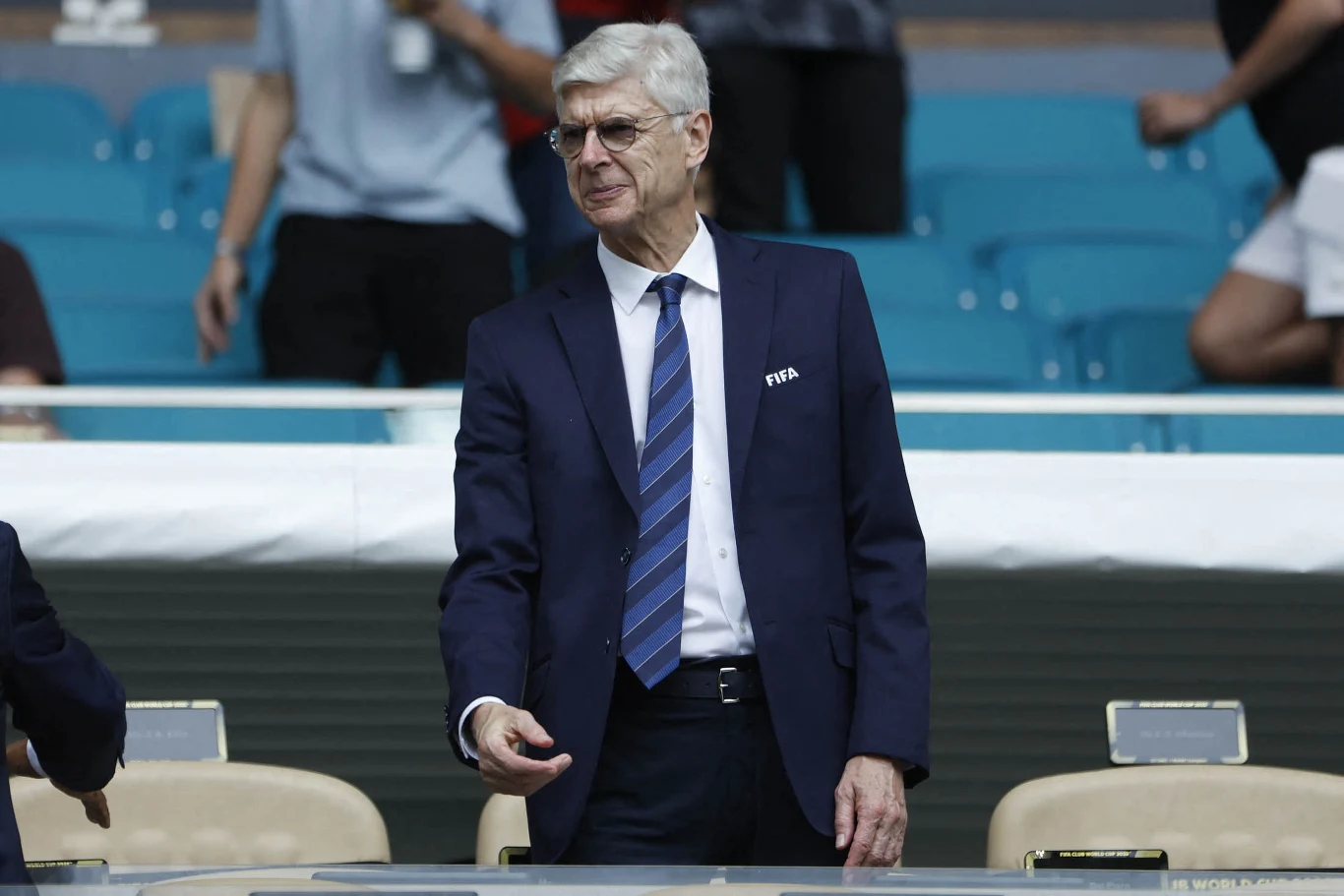 Starszy mężczyzna w garniturze i krawacie, z przypinką FIFA na marynarce, stoi w częściowo pustym sektorze stadionu, w tle widoczni są luźno rozmieszczeni kibice.