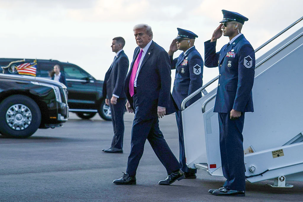 Donald Trump wysiadający z Air Force One w Palm Beach na Florydzie Mężczyzna w garniturze z różowym krawatem schodzi po schodach samolotowych na płytę lotniska, po jego prawej stronie stoi salutujący żołnierz w mundurze Sił Powietrznych, w tle widoczny samochód z amerykańską flagą oraz osoby w mundurach i garniturach.