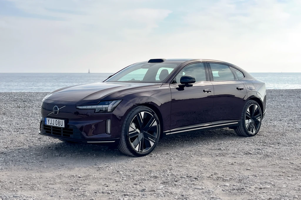 Czarny, nowoczesny samochód osobowy marki Volvo stoi zaparkowany na kamienistej plaży, w tle rozciąga się spokojne morze i jasne niebo.