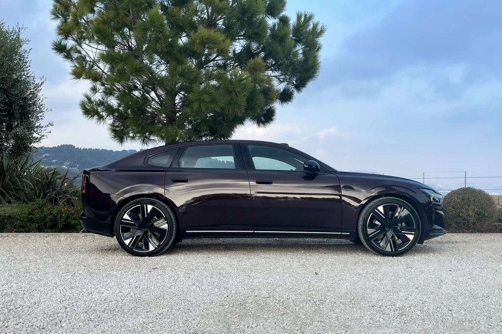 Ciemnofioletowy sedan klasy premium zaparkowany na żwirowym podjeździe na tle zielonego drzewa, z górzystym krajobrazem i niebieskim niebem w tle.