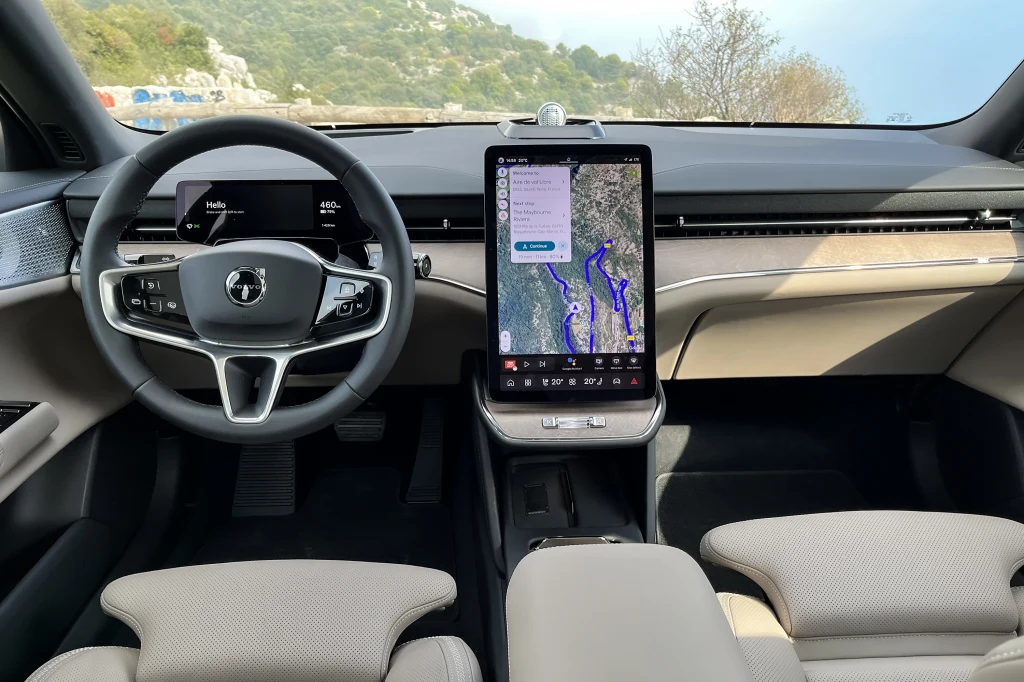 Nowoczesne wnętrze samochodu z dużym wyświetlaczem centralnym, na którym widoczna jest nawigacja, kierownica z logo producenta oraz elegancka deska rozdzielcza wykończona jasnymi i ciemnymi materiałami.