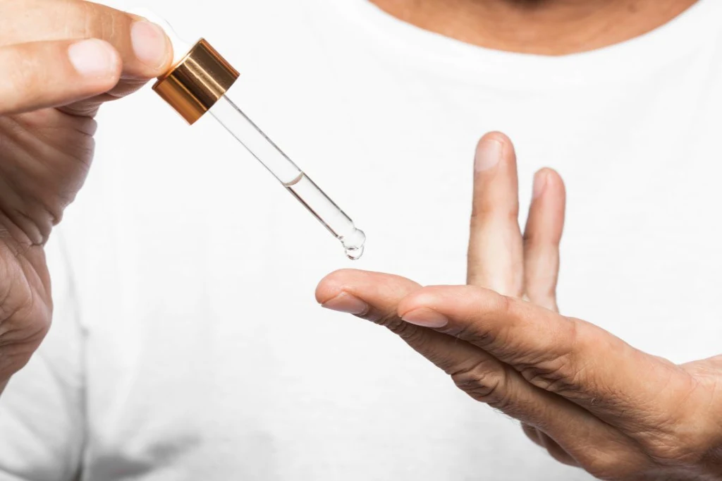 Serum, maseczka, krem - w tych kosmetykach można w Polsce znaleźć egzosomy Ręka trzymająca pipetę z przezroczystym płynem nad palcami drugiej dłoni, przygotowując się do nałożenia kropli na skórę.