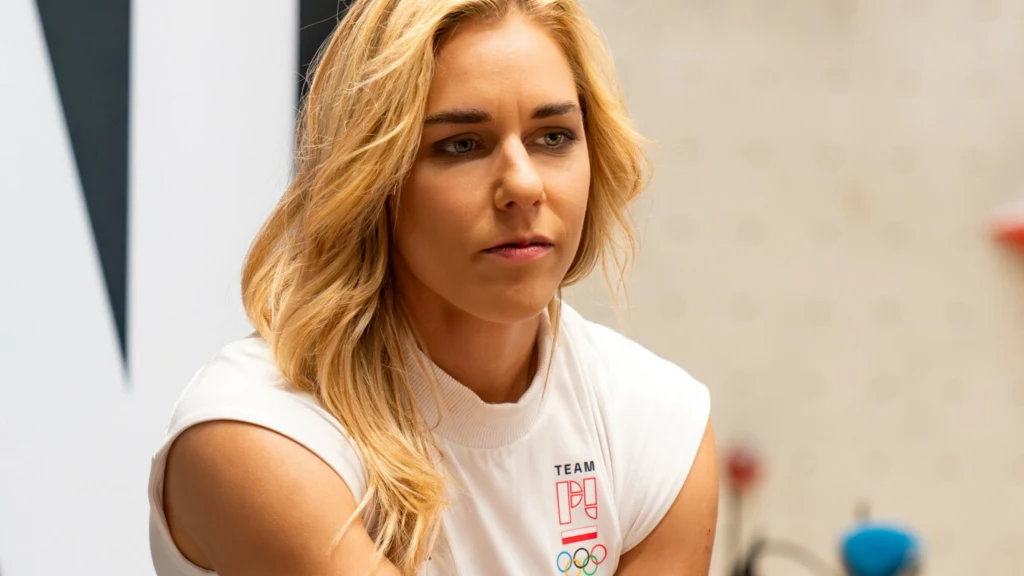Kobieta o blond włosach w białej koszulce sportowej z logo polskiej drużyny olimpijskiej, siedząca w nowoczesnym wnętrzu, skoncentrowana i zamyślona.