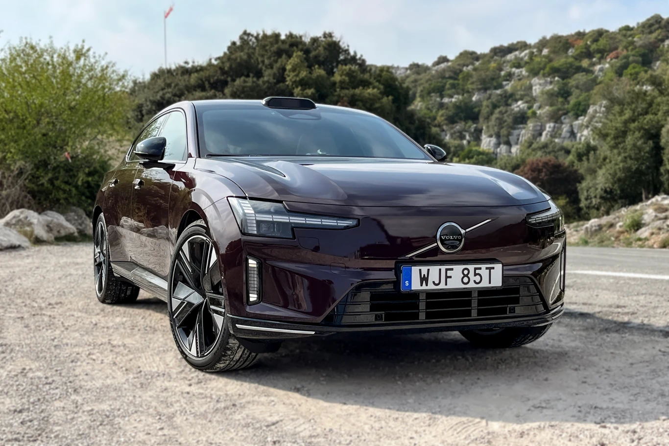 Ciemnofioletowy samochód marki Volvo o nowoczesnym, sportowym designie zaparkowany na poboczu drogi. W tle widoczna roślinność, krzewy oraz skaliste wzgórza.