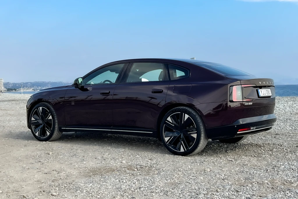 Nowoczesny, ciemnofioletowy sedan marki Polestar 2 zaparkowany na kamienistej plaży z widokiem na błękitne niebo i linię brzegową w tle.