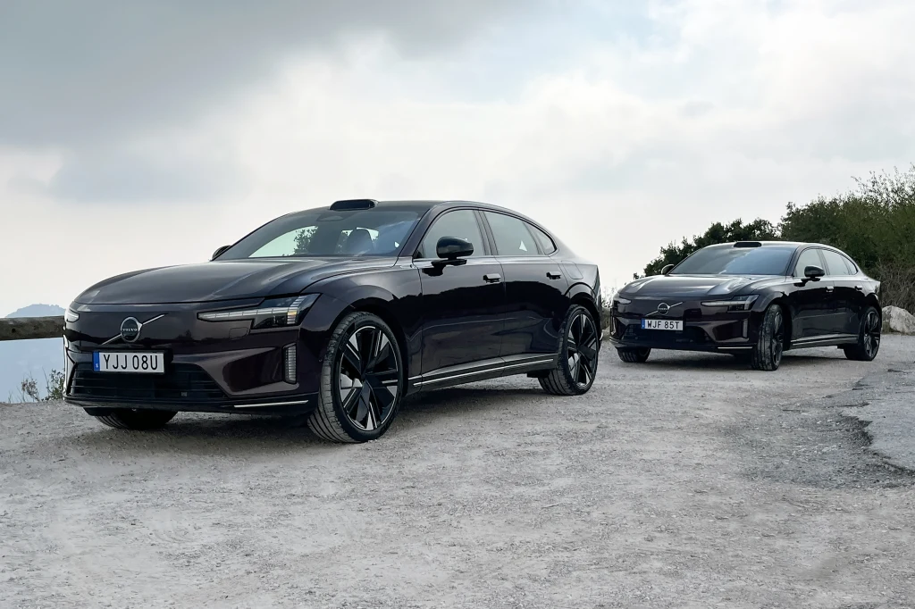 Dwa czarne samochody elektryczne klasy premium stoją zaparkowane na szutrowej drodze na tle lekko pochmurnego nieba, na pierwszym planie widoczny jest nowoczesny sedan Volvo o dynamicznej sylwetce.