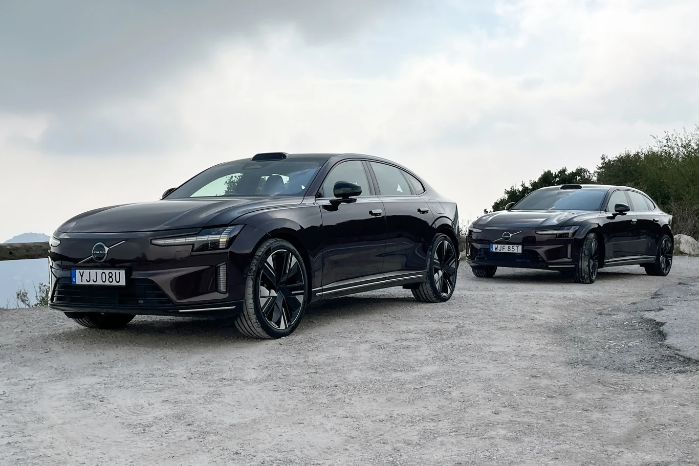 Dwa czarne samochody elektryczne klasy premium stoją zaparkowane na szutrowej drodze na tle lekko pochmurnego nieba, na pierwszym planie widoczny jest nowoczesny sedan Volvo o dynamicznej sylwetce.