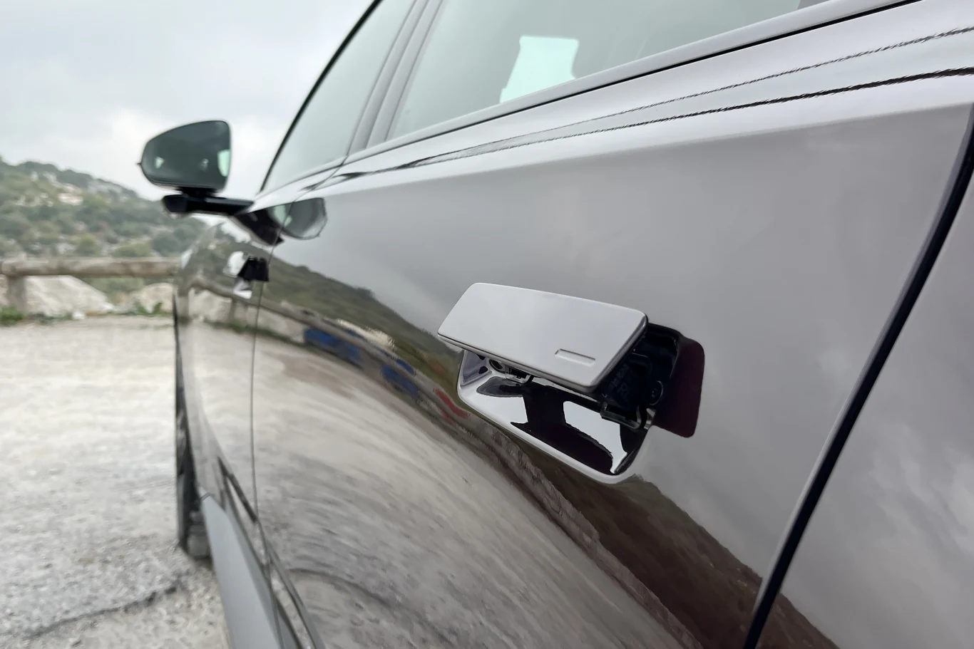 Nowoczesna klamka samochodowa o wysuwanej konstrukcji umieszczona na błyszczących drzwiach auta, widoczna z bliska, w tle rozmyty krajobraz oraz fragment lusterka bocznego.