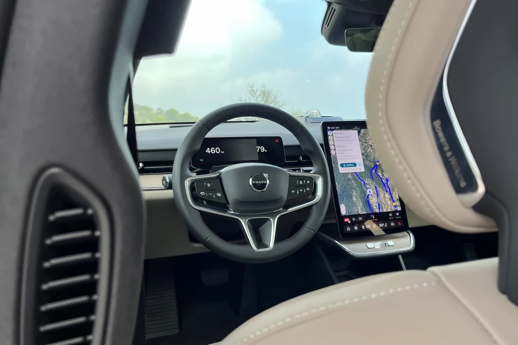 Nowoczesne wnętrze samochodu z cyfrowym kokpitem, dwuramienną kierownicą z logo marki Volvo oraz dużym dotykowym ekranem systemu multimedialnego z nawigacją, widoczne skórzane elementy fotela.