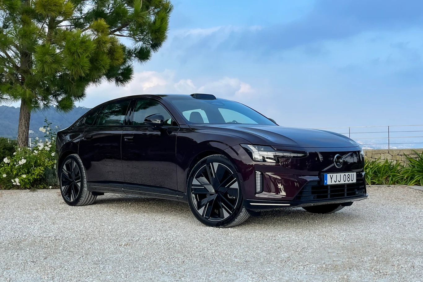 Ciemnofioletowy samochód elektryczny Polestar 2 stoi na żwirowej nawierzchni, w tle widoczne są rośliny ozdobne oraz fragment metalowego płotu, z lewej strony rozłożyste drzewo.