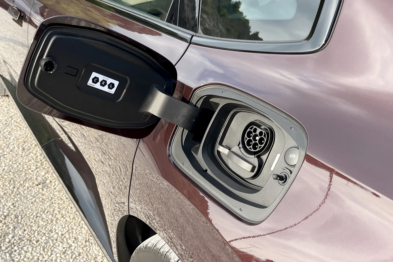 Otwarte gniazdo ładowania w samochodzie elektrycznym, zamontowane na ciemnym nadwoziu pojazdu, widoczne na tle drogi lub parkingu.