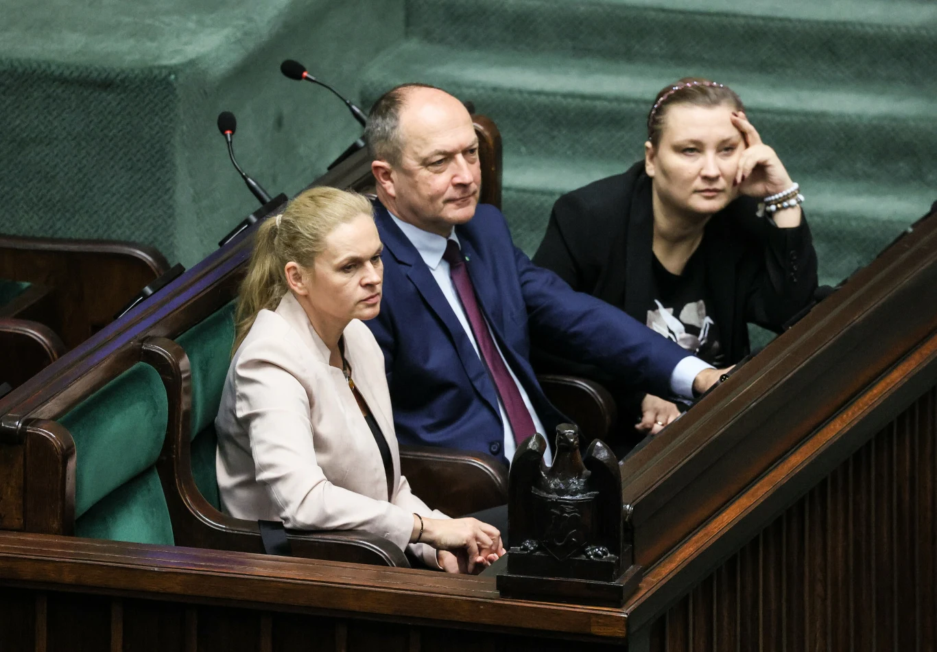 Gotowi do pracy, ale bez pensji. Nowe przepisy uderzają w nauczycieli (zdj. ilustracyjne) Trzy osoby siedzące w ławach sejmowych, skupione i zamyślone, mężczyzna w garniturze z dwoma kobietami po bokach; w tle klasyczne wnętrze izby parlamentarnej.