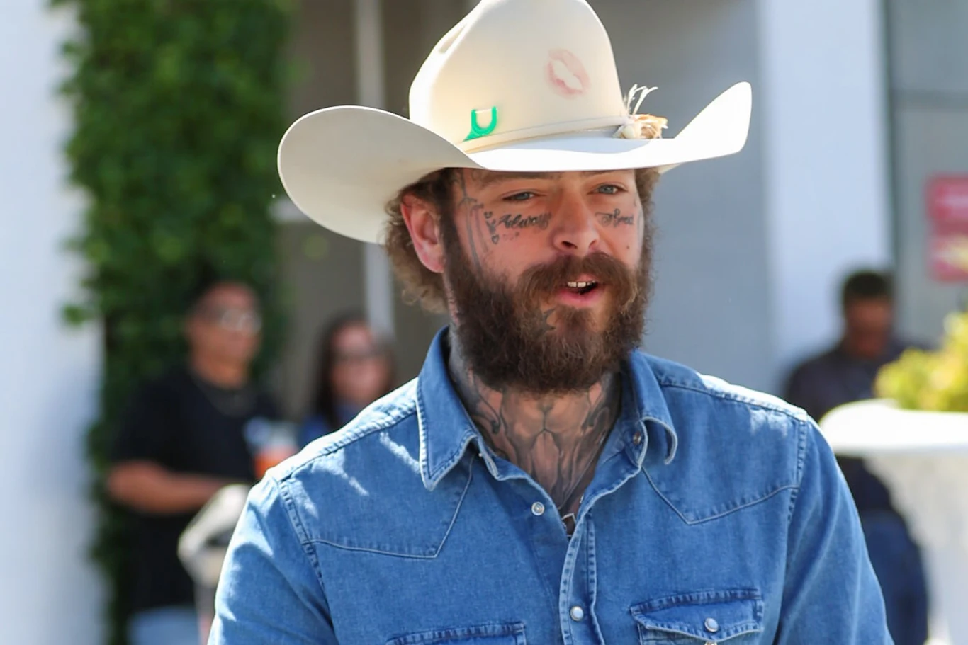 Post Malone został pozwany przez kierowcę, który wystąpił w jego teledysku Mężczyzna z brodą i tatuażami na twarzy, ubrany w niebieską koszulową kurtkę i biały kowbojski kapelusz, stoi na zewnątrz. W tle rozmyte postacie oraz fragment zieleni i budynku.