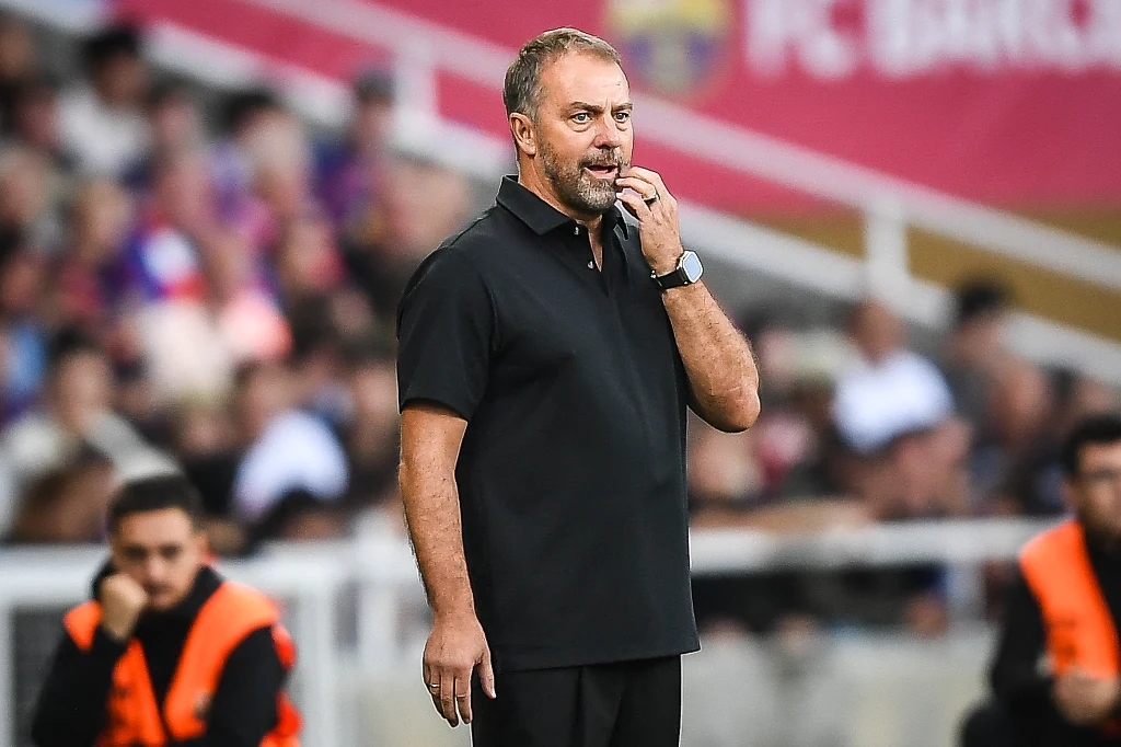 Mężczyzna ubrany w czarną koszulkę polo stoi przy linii bocznej boiska podczas meczu piłkarskiego, skupiony na grze, w tle rozmyty tłum kibiców oraz zabezpieczenie techniczne.