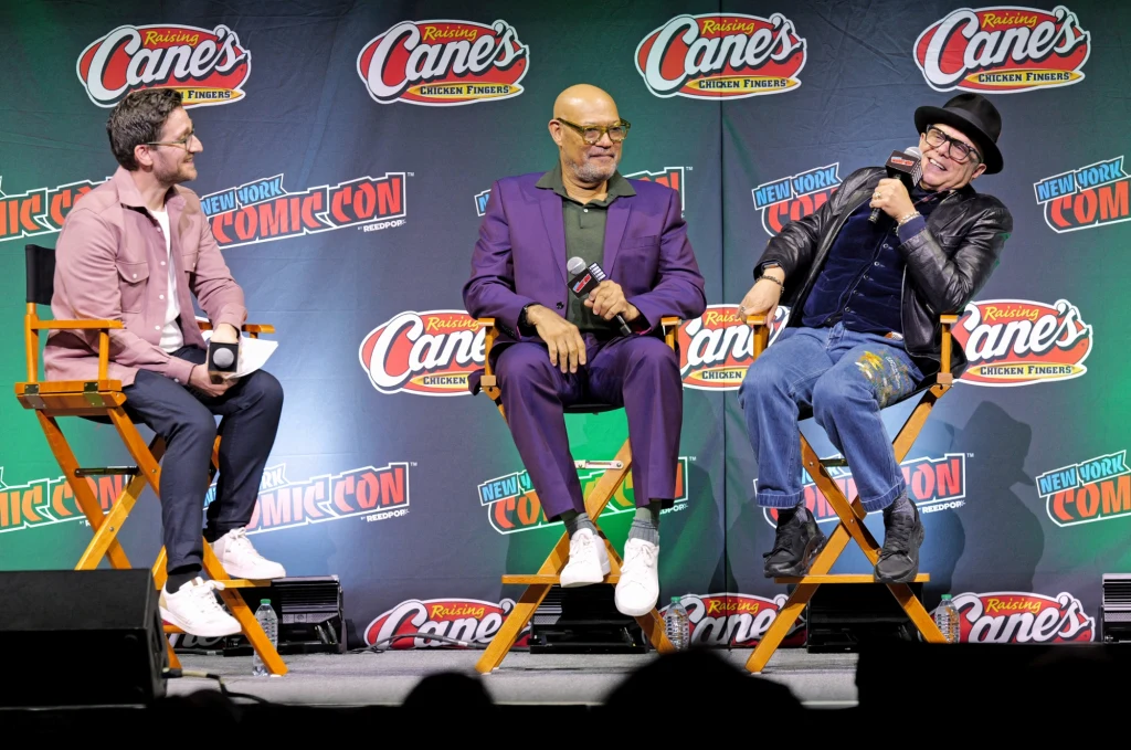 Trzech mężczyzn siedzi na wysokich żółtych krzesłach na scenie podczas panelu dyskusyjnego na tle banera promocyjnego z logo Raising Cane's oraz New York Comic Con, dwóch z nich trzyma mikrofony, jeden gestykuluje i śmieje się.