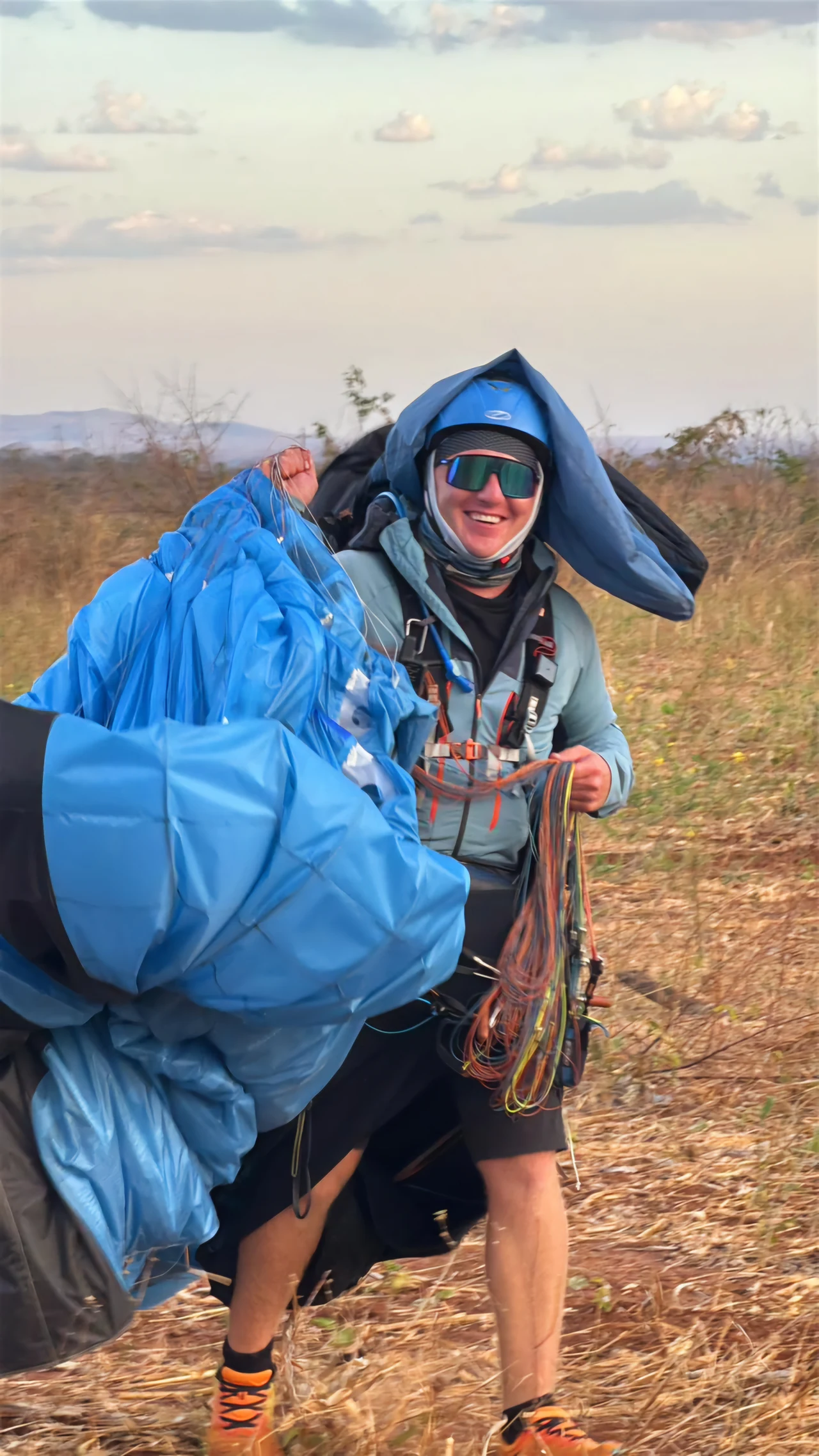 Mateusz Siatka Uśmiechnięty mężczyzna w okularach przeciwsłonecznych z paralotnią i sprzętem na suchym, trawiastym terenie, ubrany sportowo, z kapturem na głowie, w otoczeniu przyrody pod lekko zachmurzonym niebem.