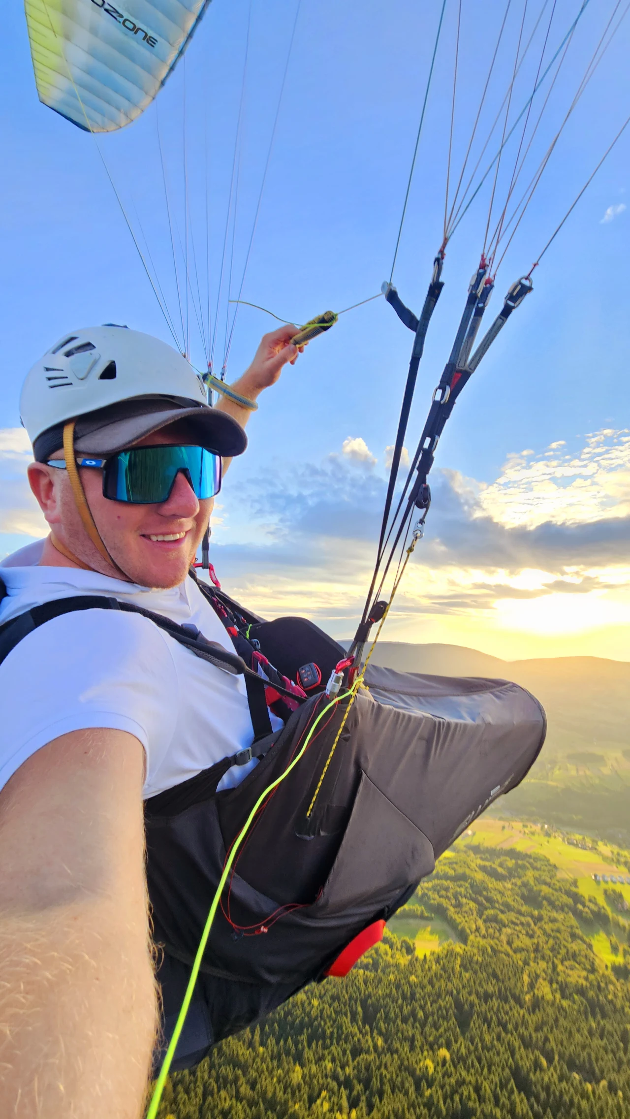Mateusz Siatka Mężczyzna w kasku i okularach przeciwsłonecznych podczas lotu paralotnią nad zielonymi polami i lasami oświetlonymi słońcem, na tle malowniczego krajobrazu i błękitnego nieba z chmurami.