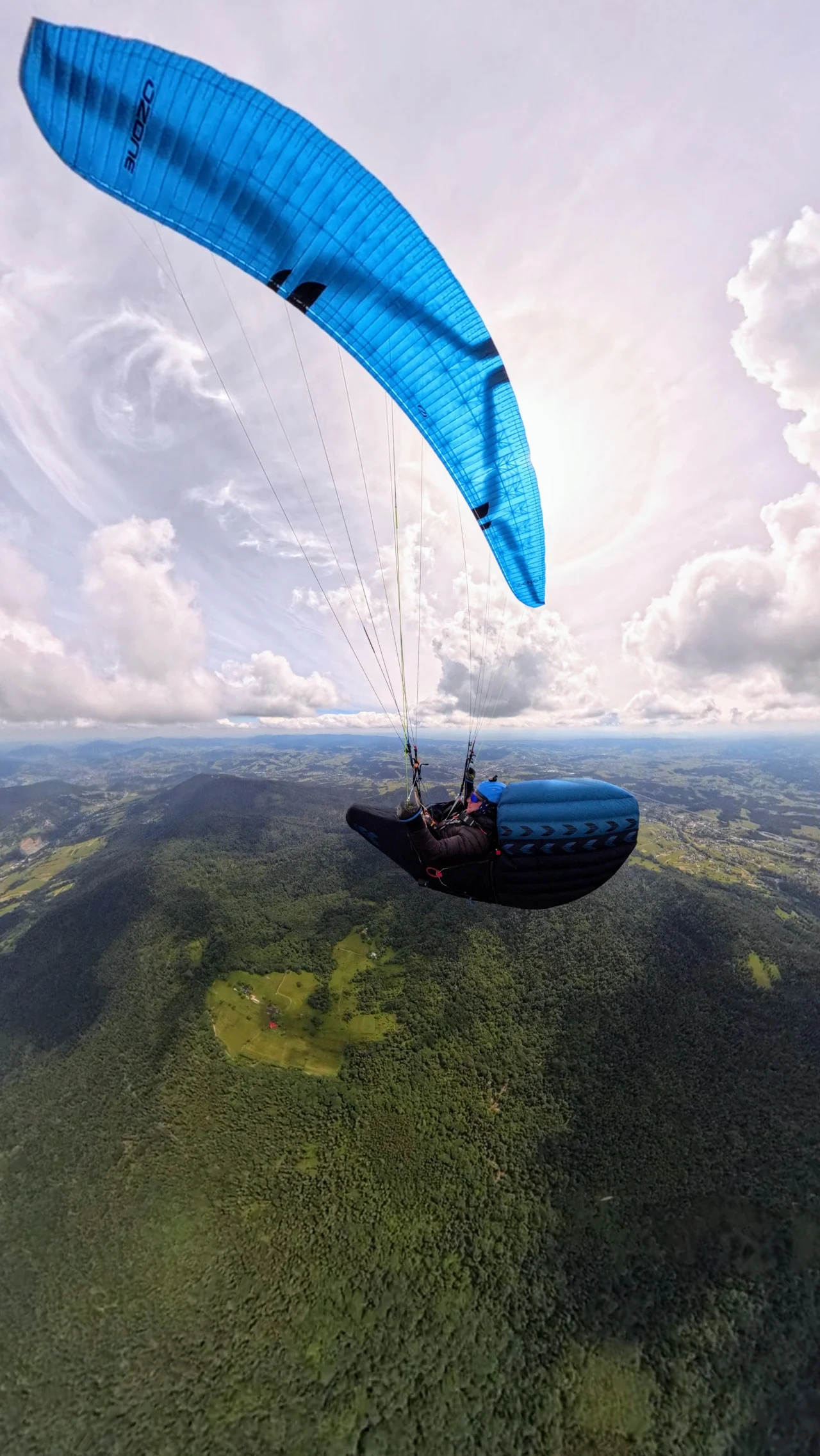 Mateusz Siatka Paralotniarz unoszący się wysoko nad zielonymi lasami i polami, pod błękitnym skrzydłem na tle rozległego nieba z dynamicznymi chmurami