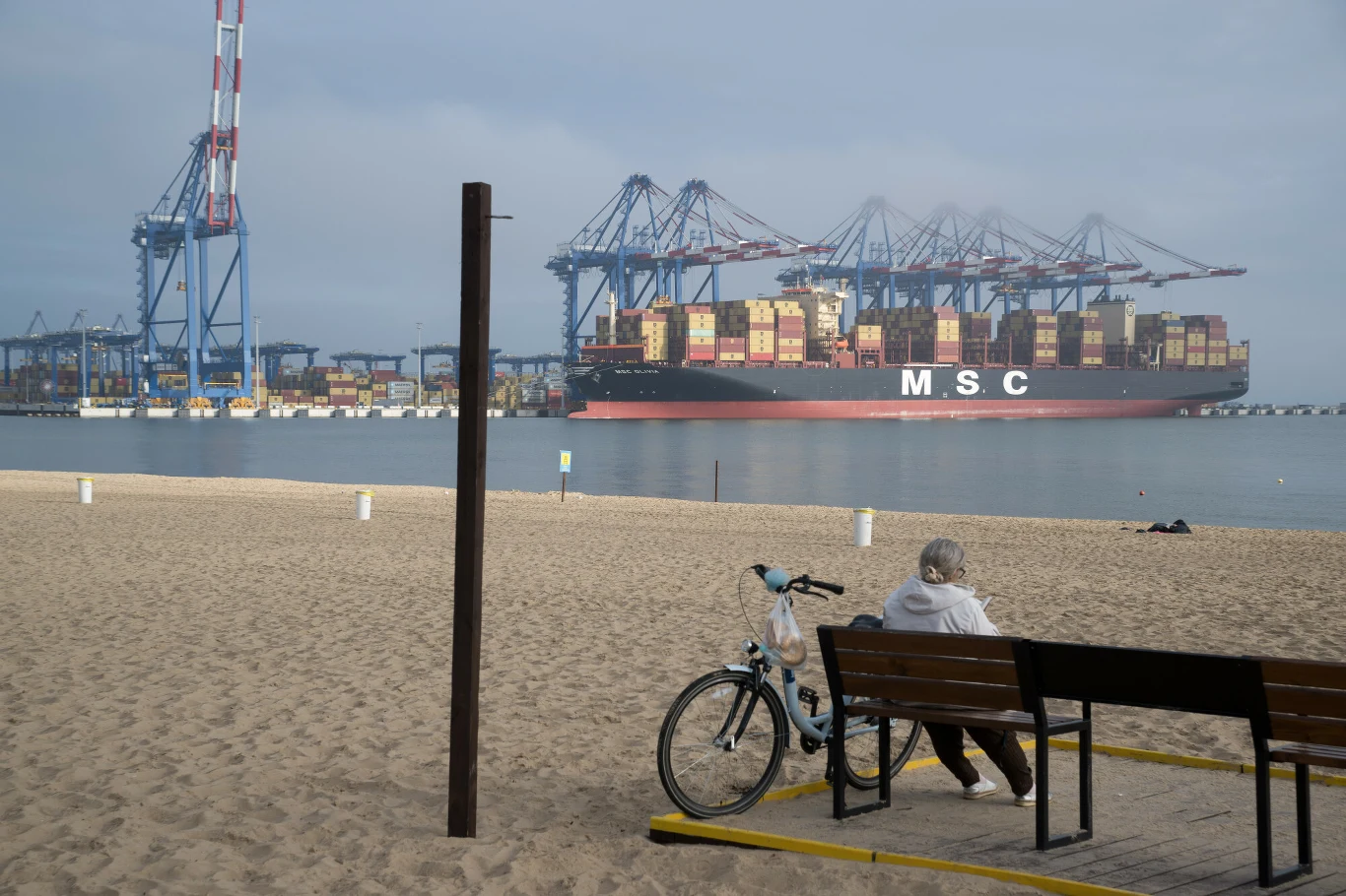 Przenoszenie produkcji do odległych i tańszych krajów nie wyszło Zachodowi na dobre. Wciąż jednak do Europy i USA płyną kontenerowce pełne towarów wyprodukowanych tysiące kilometrów od amerykańskich i europejskich miast (zdj. ilustracyjne) Statek towarowy z widocznymi kontenerami płynie w tle po wodach portu, z przodu osoba siedzi na ławce przy plaży obok roweru, na drugim planie widoczne są suwnice przeładunkowe.